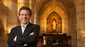 Professor Anthony Kronman stands with arms crossed in a large stone-walled room with vaulted ceiling and the Alma Mater portrait, a painting of a standing woman, in the distance